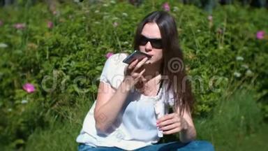 阳光明媚的一天，美丽的黑发女人<strong>坐在公园里</strong>，在手机上记录了一条语音信息。
